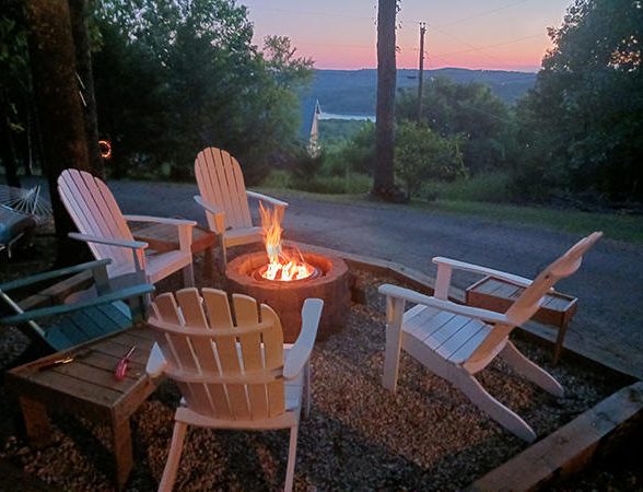 Four white Adirondack chairs around a glowing fire pit at dusk with trees and lake view at Foxfire Resort On Table Rock Lake